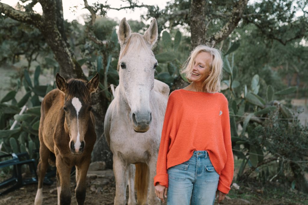 FRau lachend neben weißem Pferd und Fohlen. Sinnbild für pferdegestütztes Coaching und Neubeginn